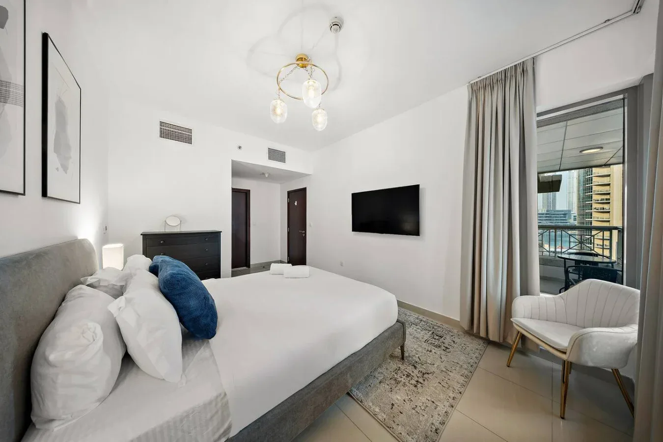 Un dormitorio luminoso con una cama grande, un televisor en la pared y cortinas junto a una puerta de vidrio.