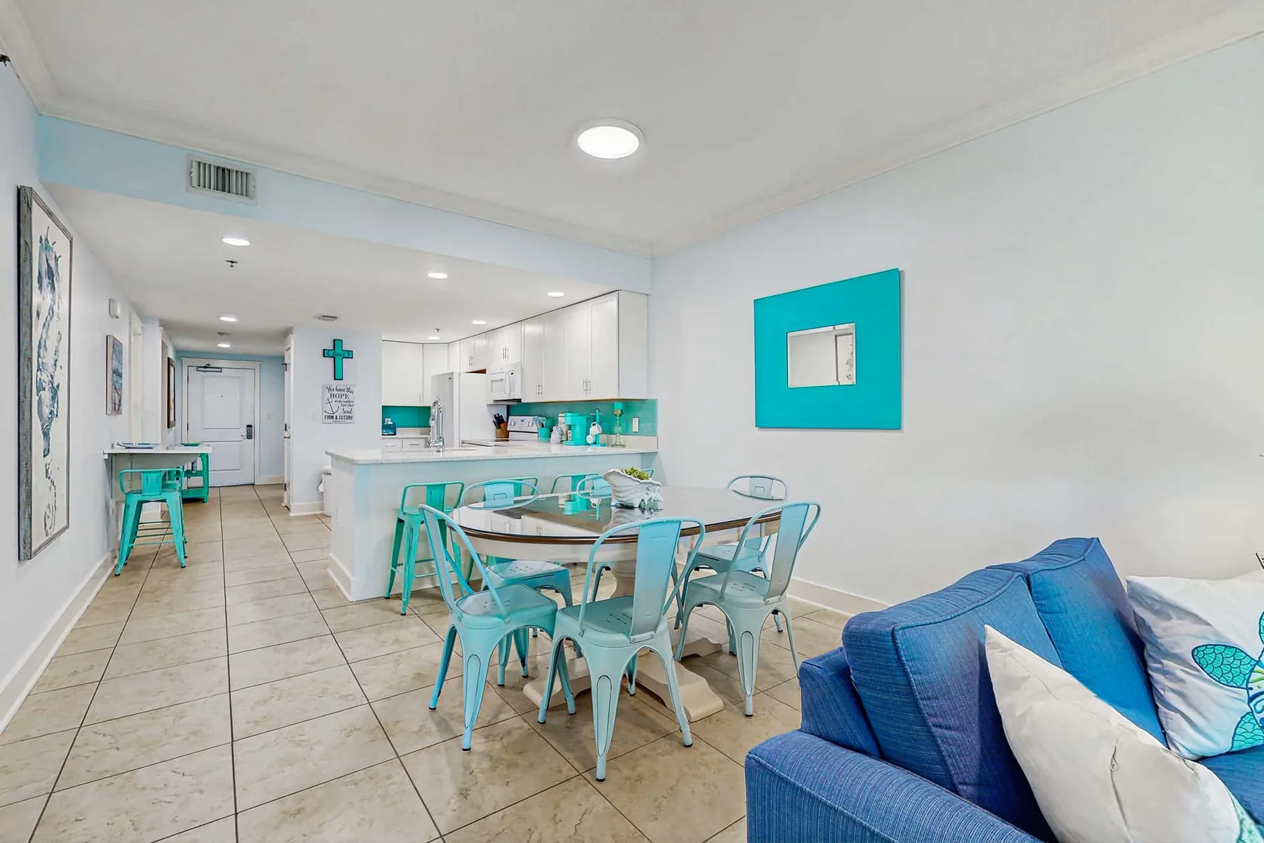 A bright dining area with a glass table beside a turquoise-accented kitchen.
