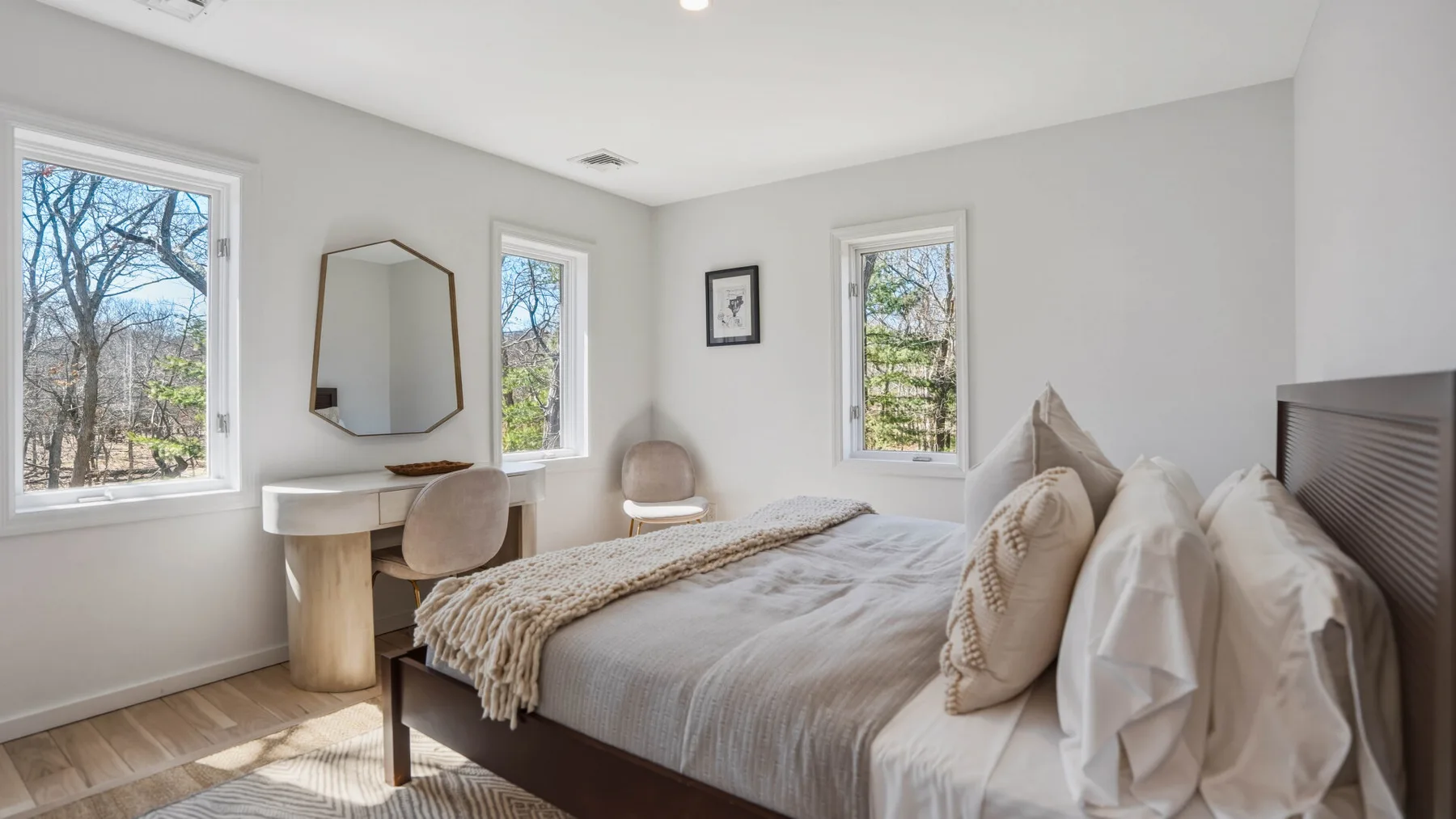 Un dormitorio luminoso con cama, tocador y ventanas con vistas a los árboles.