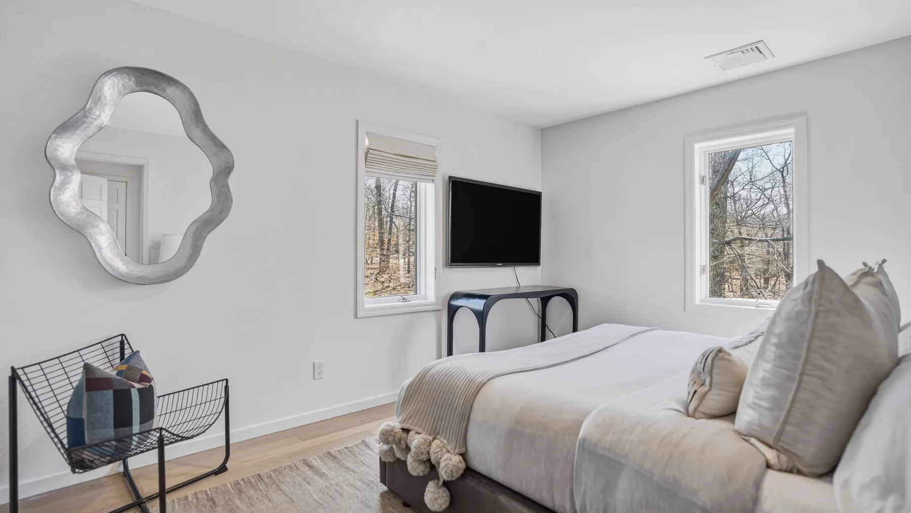 Un dormitorio luminoso con una cama, un televisor en la pared y ventanas con vistas a los árboles.