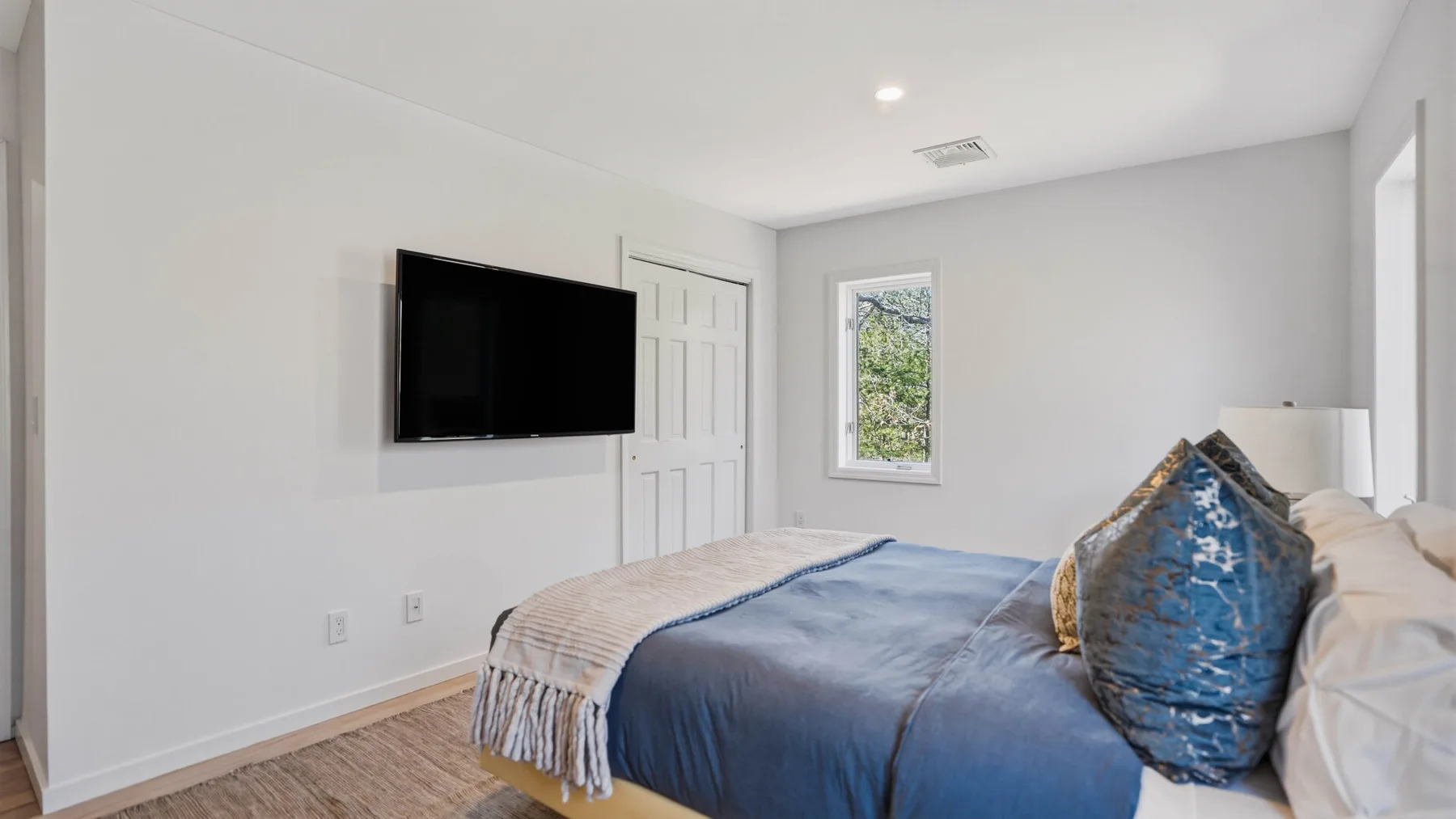 Un dormitorio luminoso con una cama, un televisor en la pared y una ventana.