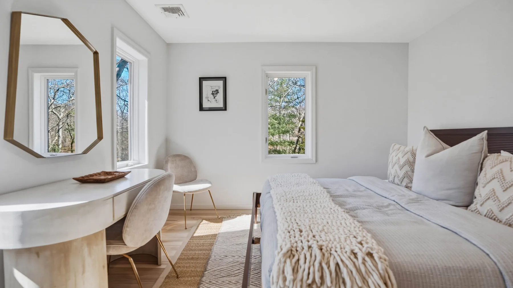 Un dormitorio luminoso con cama, una pequeña zona de estar y ventanas con vistas a los árboles.
