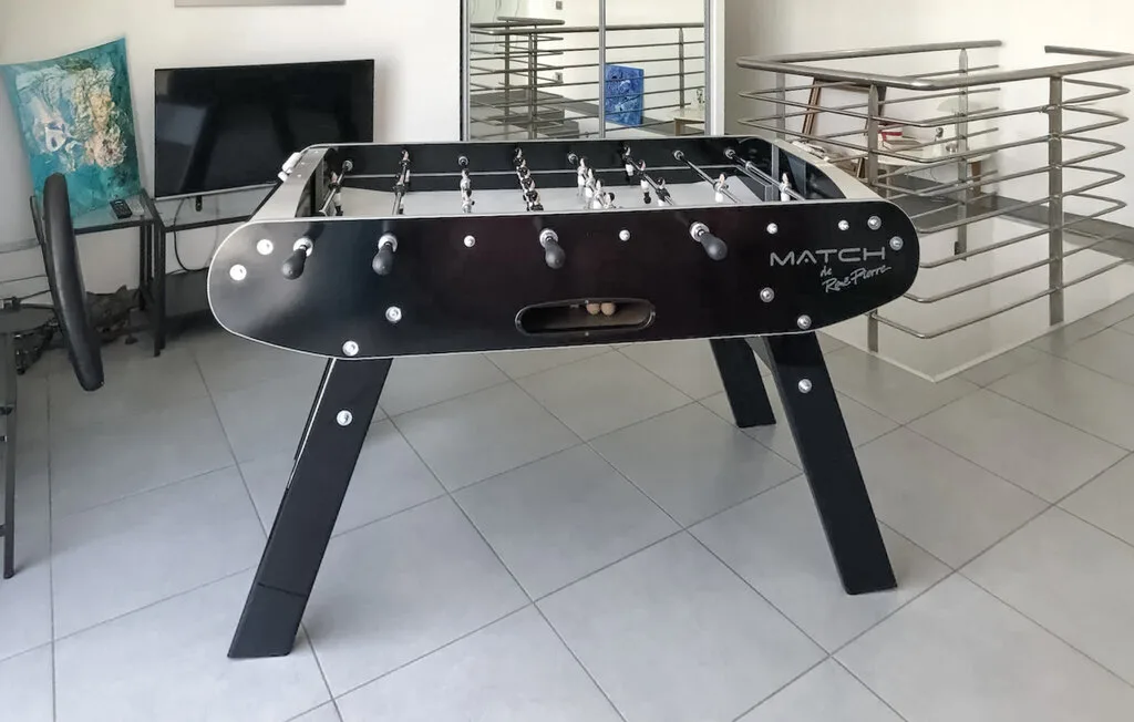 Una mesa de futbolín está en un espacio interior embaldosado junto a una barandilla metálica.