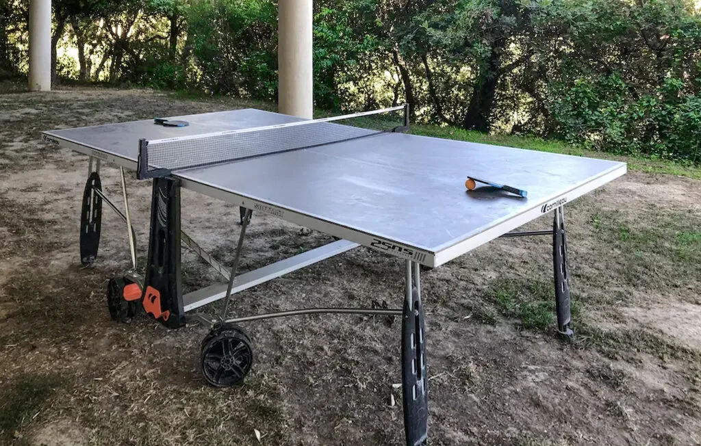 Una mesa de ping-pong al aire libre está en un patio sombreado rodeado de vegetación.