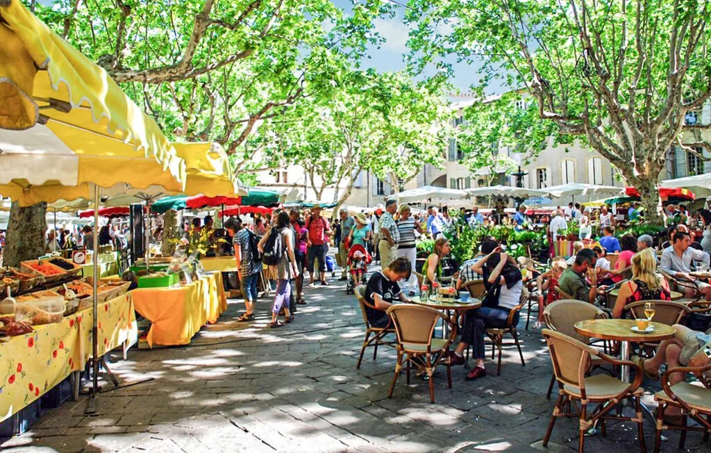 Un mercado al aire libre y un café llenan una plaza arbolada con puestos y mesas.