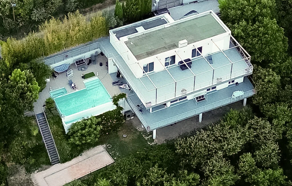 Una vista aérea muestra un edificio moderno junto a una piscina, rodeado de árboles.