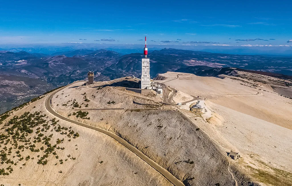 Una torre de comunicaciones se alza en una cresta montañosa árida junto a un camino sinuoso.