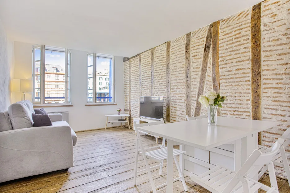 Una luminosa sala de estar y comedor con asientos blancos y una mesa junto a dos ventanas.