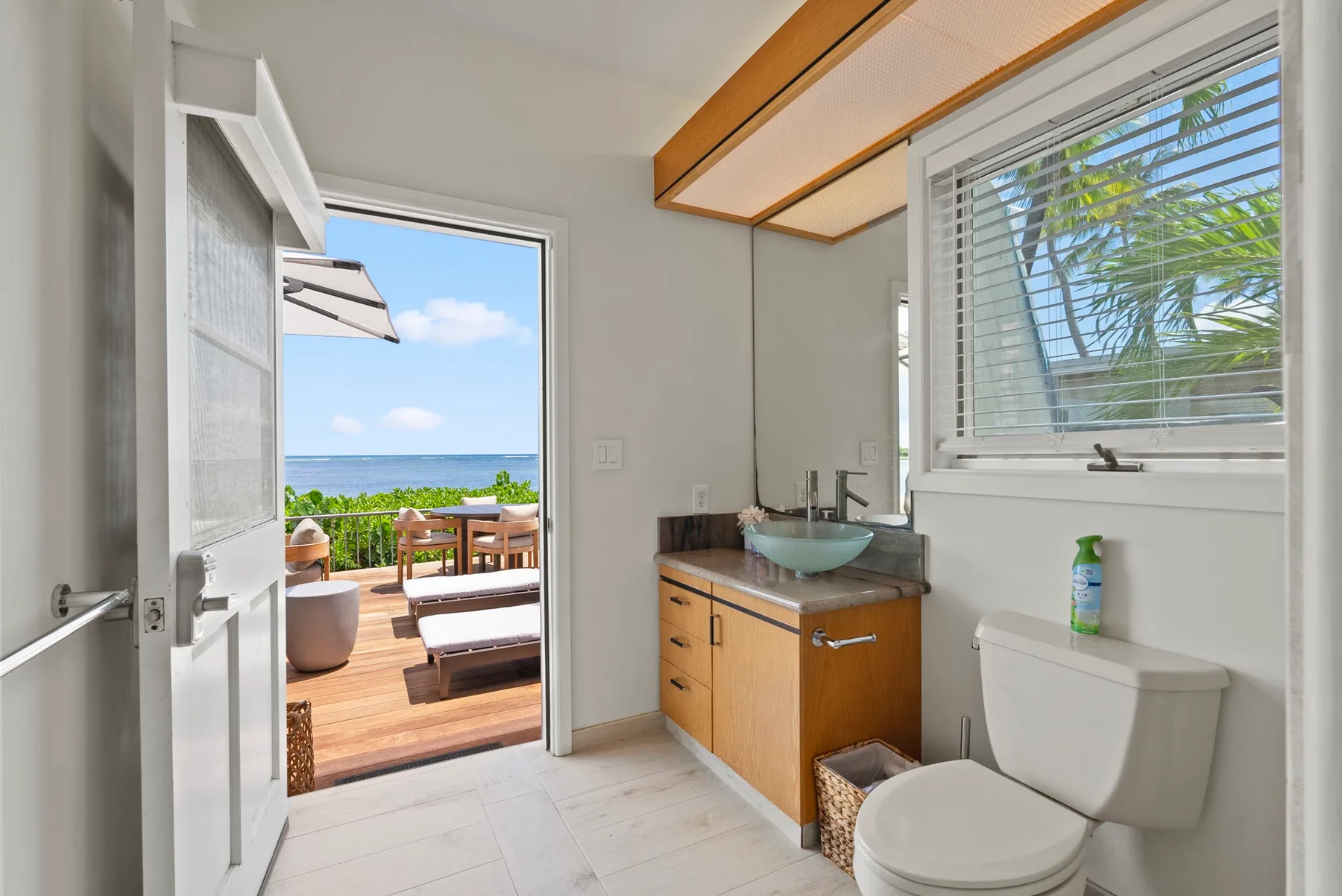 A bright bathroom opens onto a wooden deck with outdoor seating and ocean views.