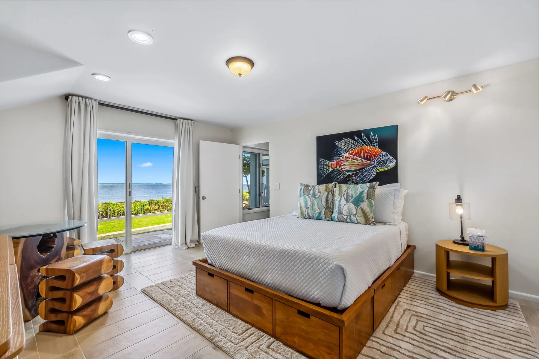 A bright bedroom features a wooden bed, colorful fish artwork, and glass doors overlooking the ocean.