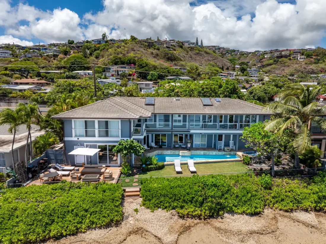 A large beachfront house overlooks a swimming pool and green hillside.