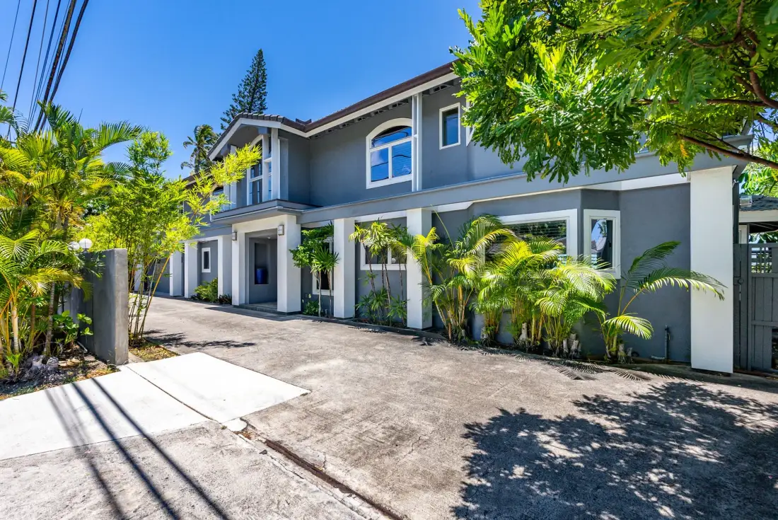 A gray multi-unit building faces a paved driveway lined with tropical plants.