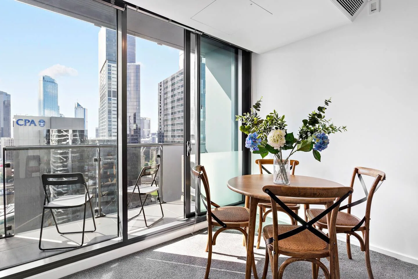 Una mesa de comedor con sillas está junto a ventanales con vistas a edificios de la ciudad.