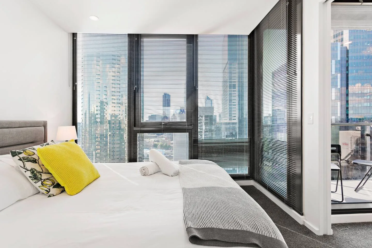 Un dormitorio con una cama junto a grandes ventanas con vistas a edificios cercanos.