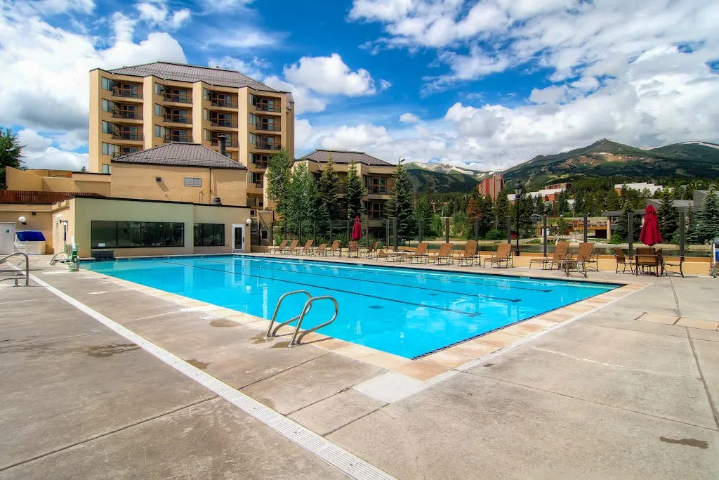 An outdoor swimming pool sits beside a hotel building, with mountains in the background.