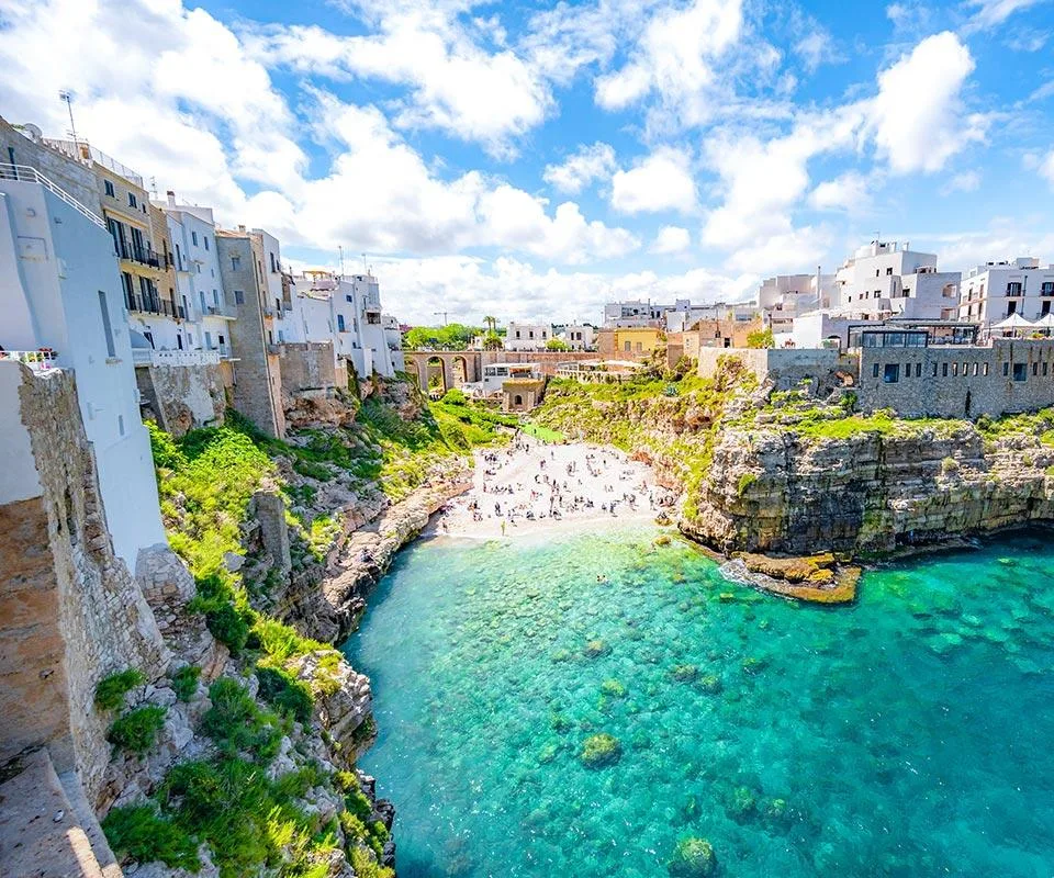 Playa costera con agua cristalina, edificios históricos y acantilados verdes.