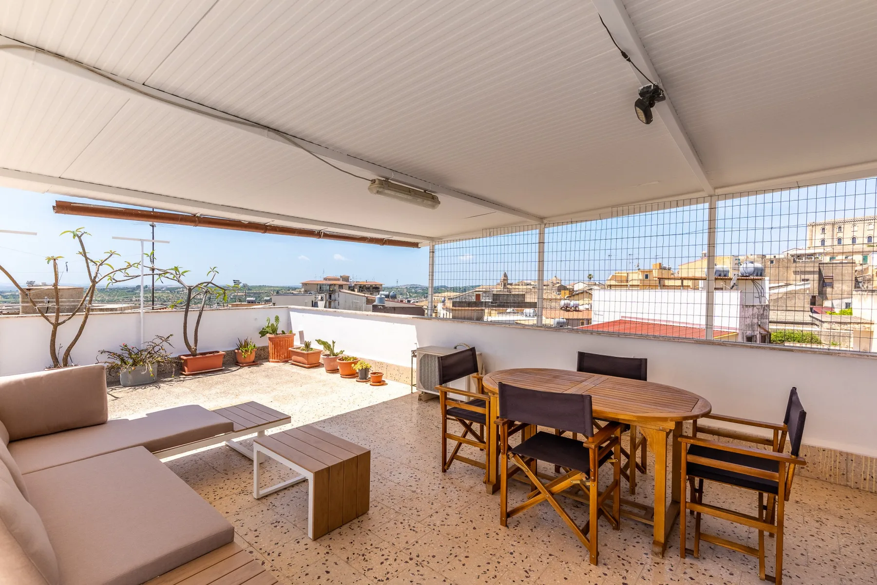 Una terraza cubierta tiene asientos al aire libre, una mesa de comedor de madera y vistas a edificios cercanos.