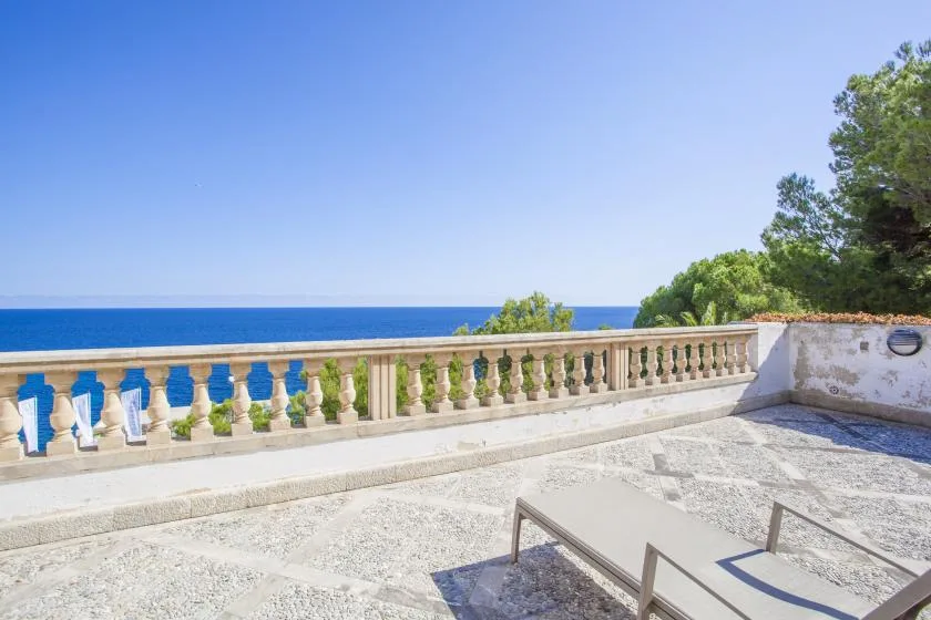 A sunlit outdoor terrace overlooks the blue sea beyond a stone balustrade.