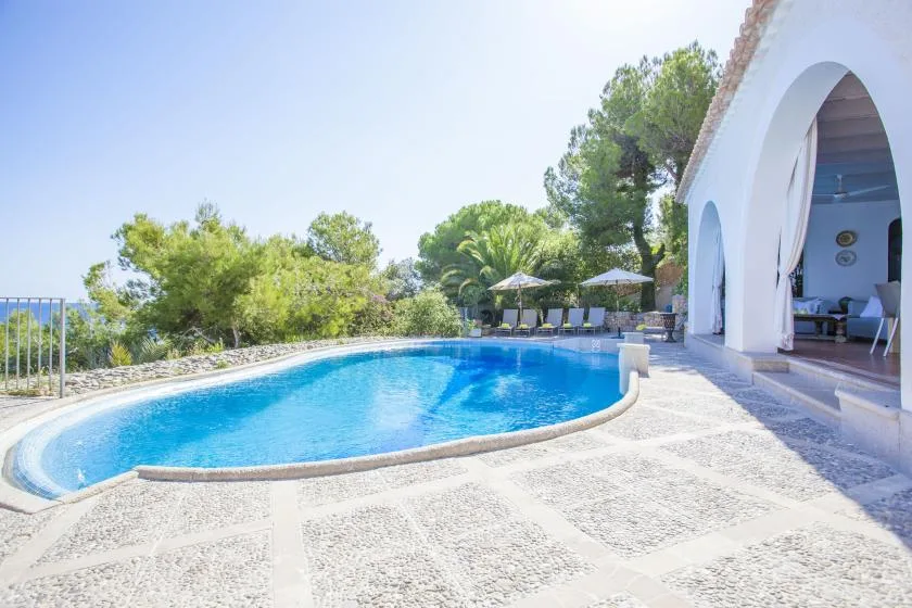 A curved outdoor swimming pool beside a white arched building and patio seating.