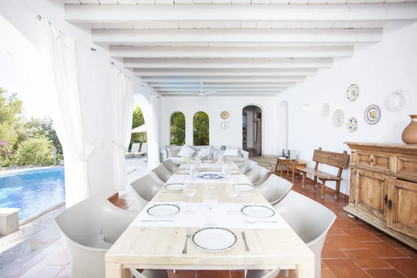 A bright dining room with a long set table beside open windows overlooking a pool.