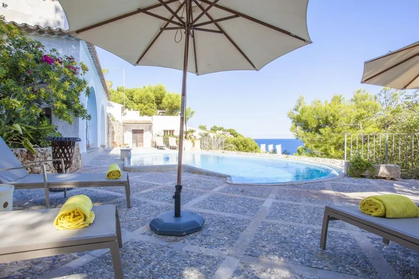 A sunny outdoor pool area with shaded seating and a view of the sea.