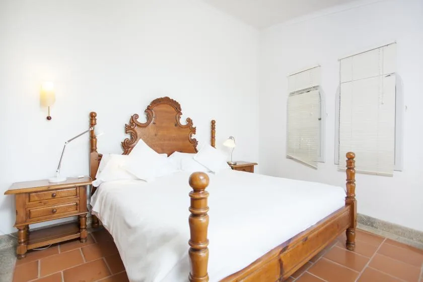 A bedroom with a wooden bed, white bedding, and windows with blinds.