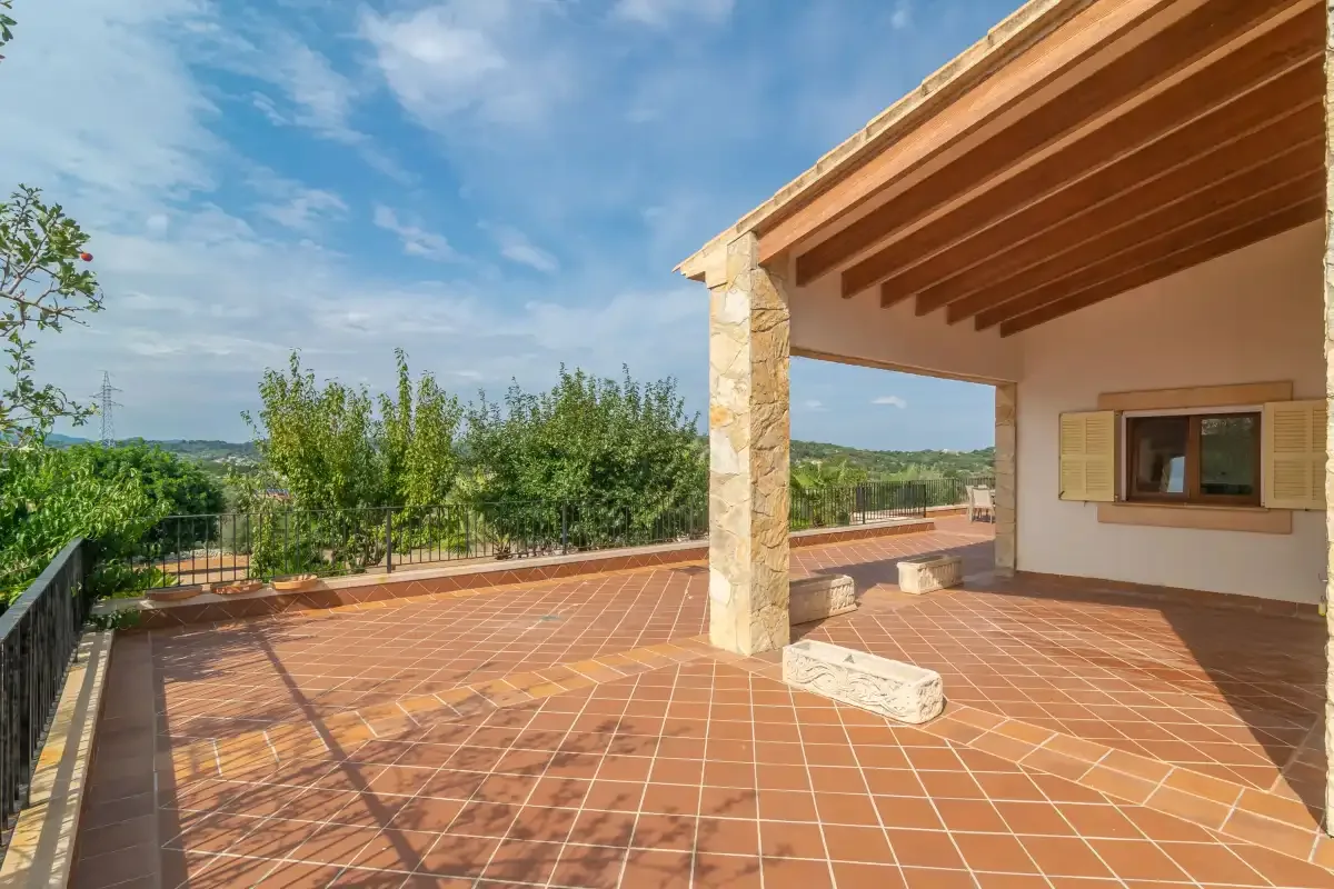 Una terraza exterior de baldosas con una zona cubierta y vistas a la vegetación.