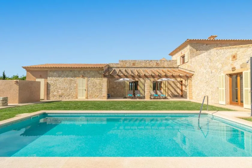 An outdoor swimming pool sits in front of a stone house and shaded patio.