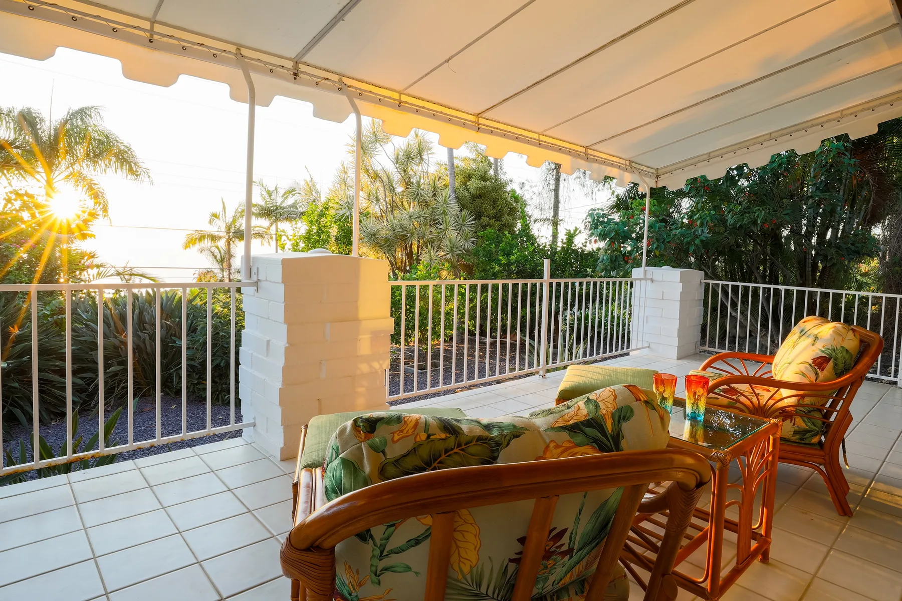 A covered patio with wicker seating and a railing overlooks palm trees.