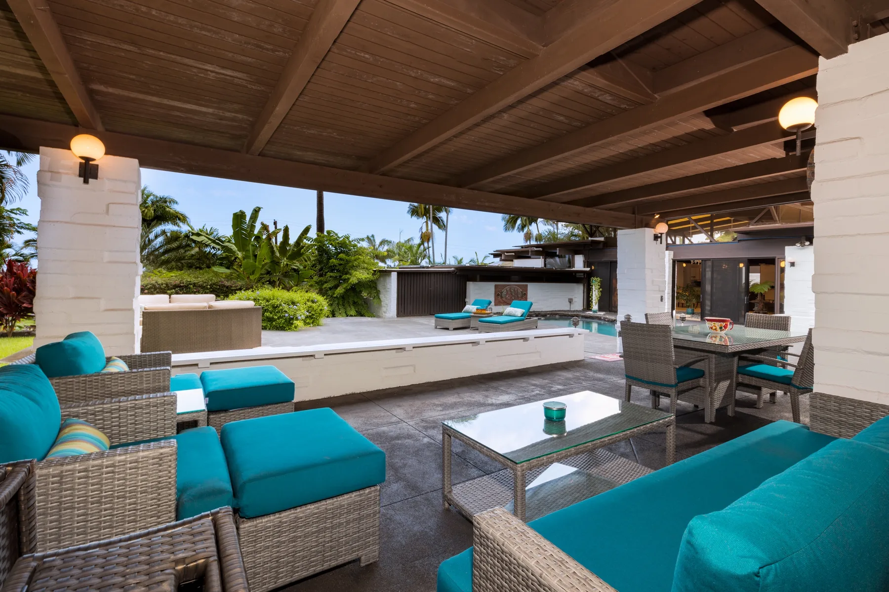 Una sala exterior cubierta con asientos turquesa da a una piscina y vegetación tropical.
