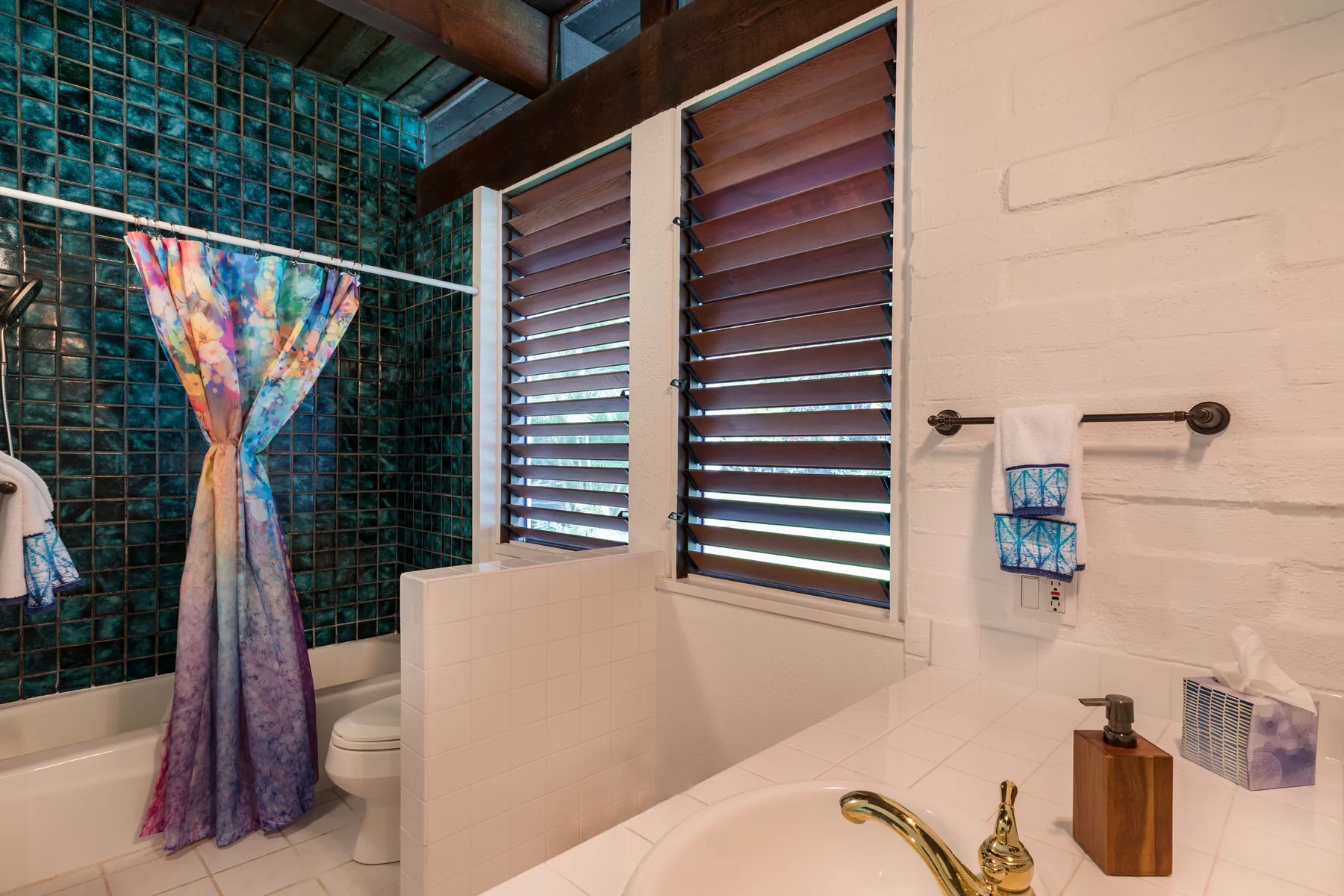 A bathroom with a bathtub, colorful shower curtain, and louvered windows.