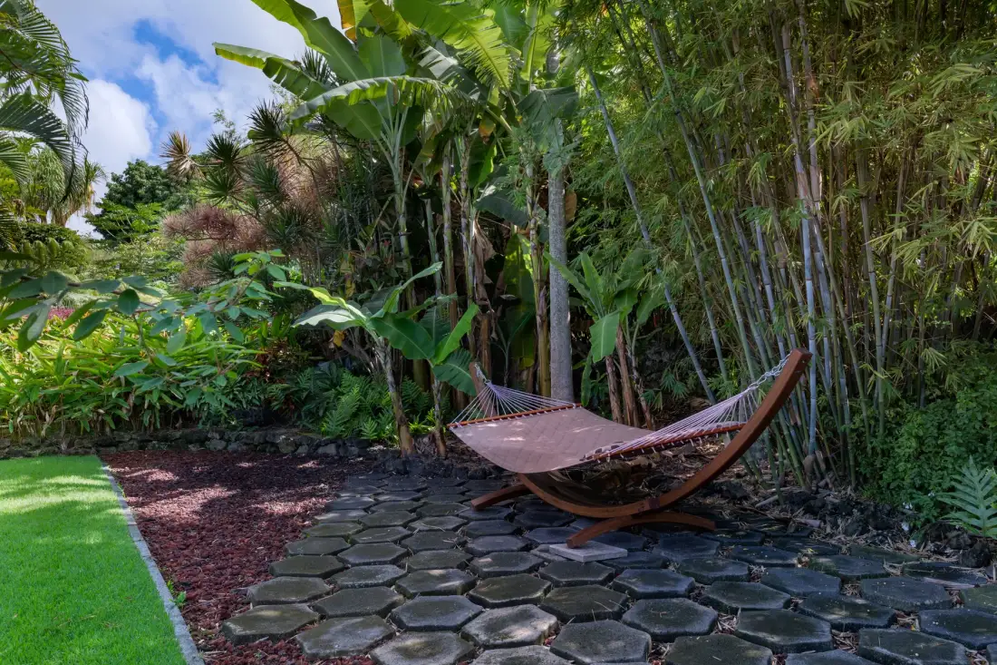 A hammock rests beside a stone path amid lush tropical garden plants.