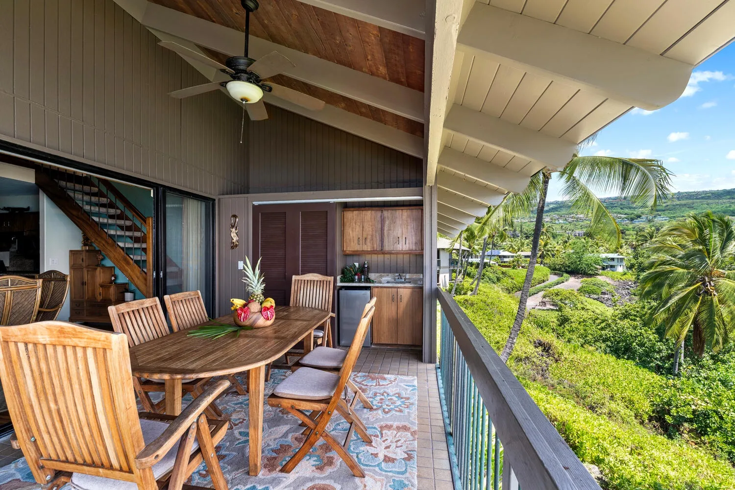 A covered outdoor dining area with a wooden table and chairs overlooks tropical greenery.