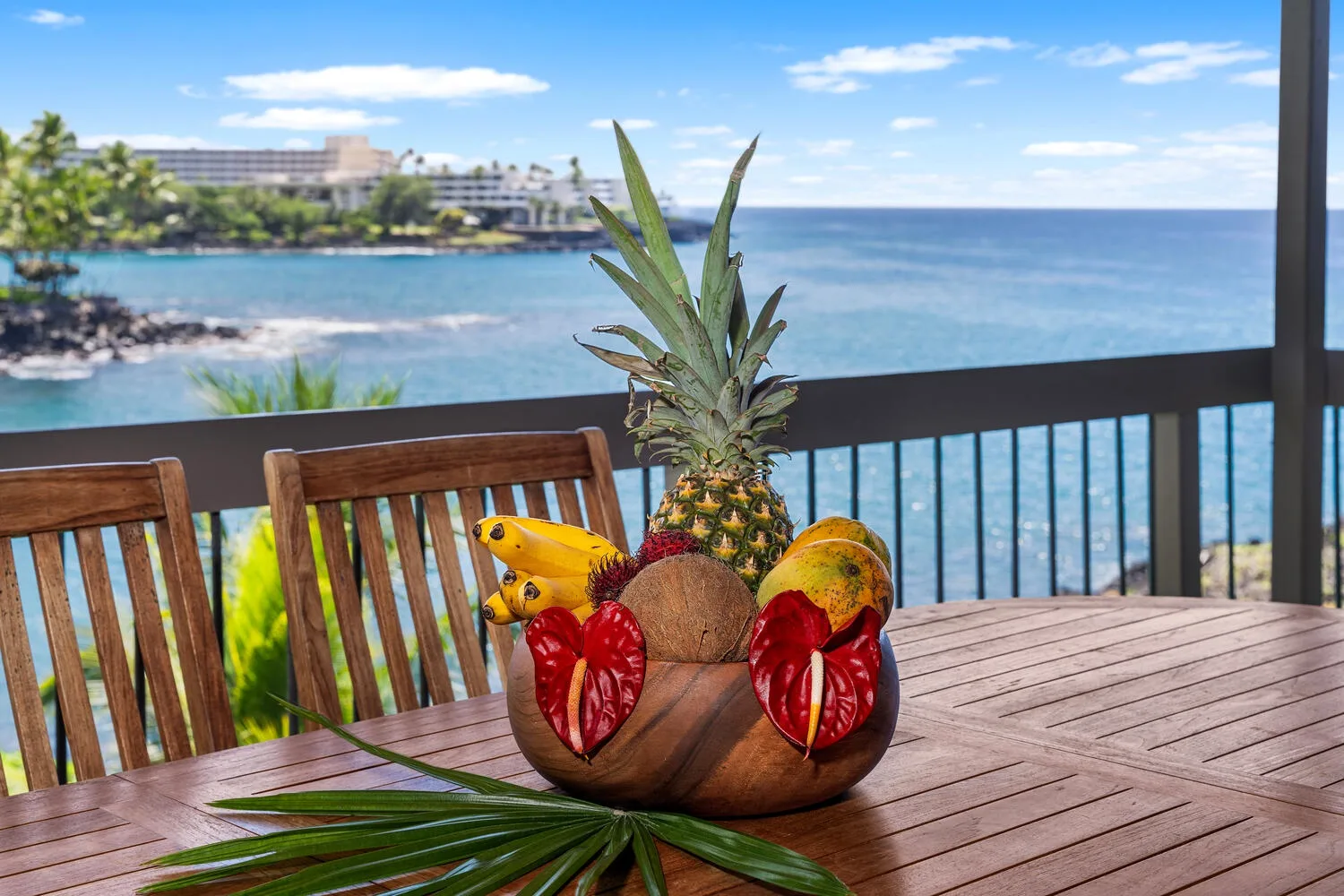 A seaside balcony table displays a pineapple and tropical fruit against an ocean view.