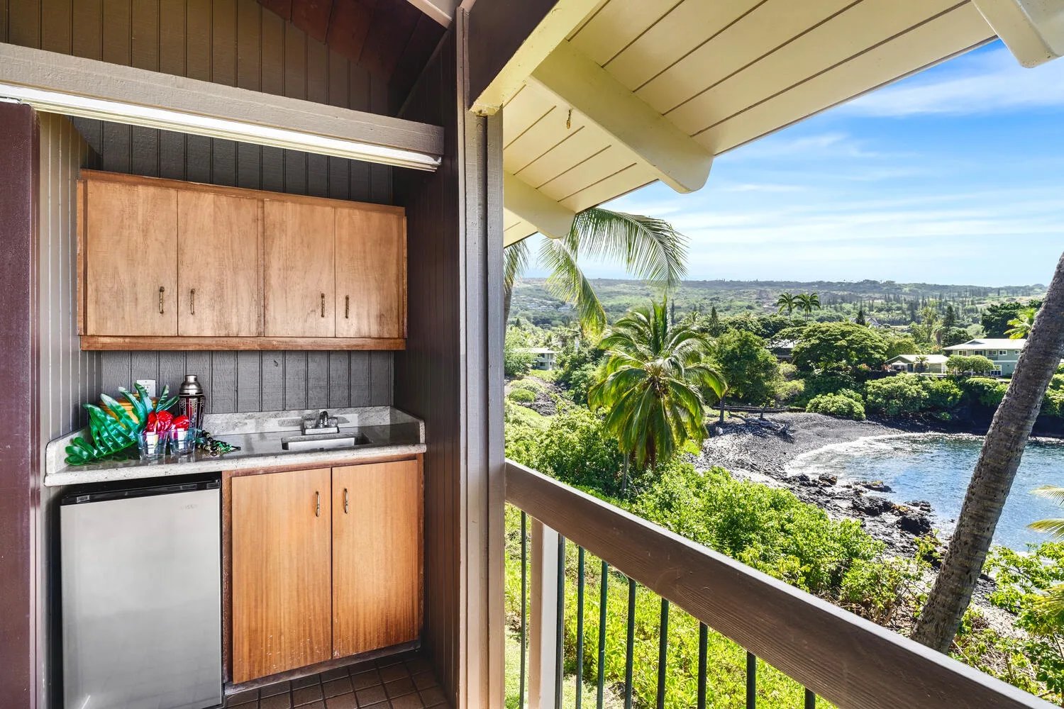 A compact kitchenette opens onto a balcony overlooking a leafy coastal landscape.