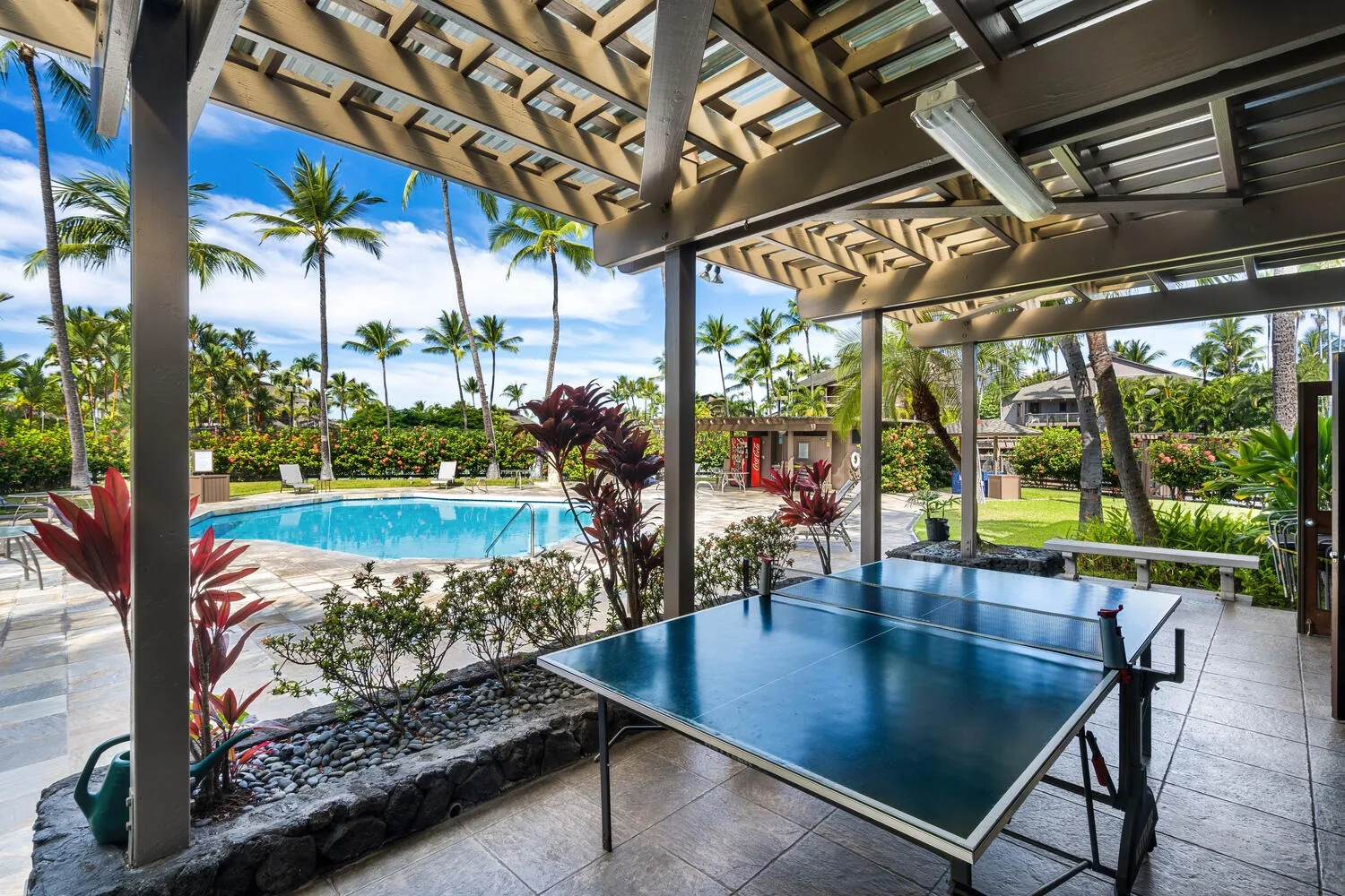 A covered patio with a ping-pong table overlooks a swimming pool and tropical landscaping.
