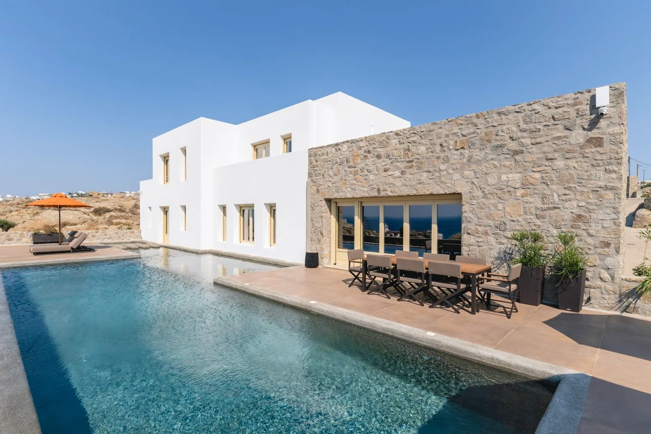 A modern villa beside a swimming pool, with outdoor seating along the stone facade.