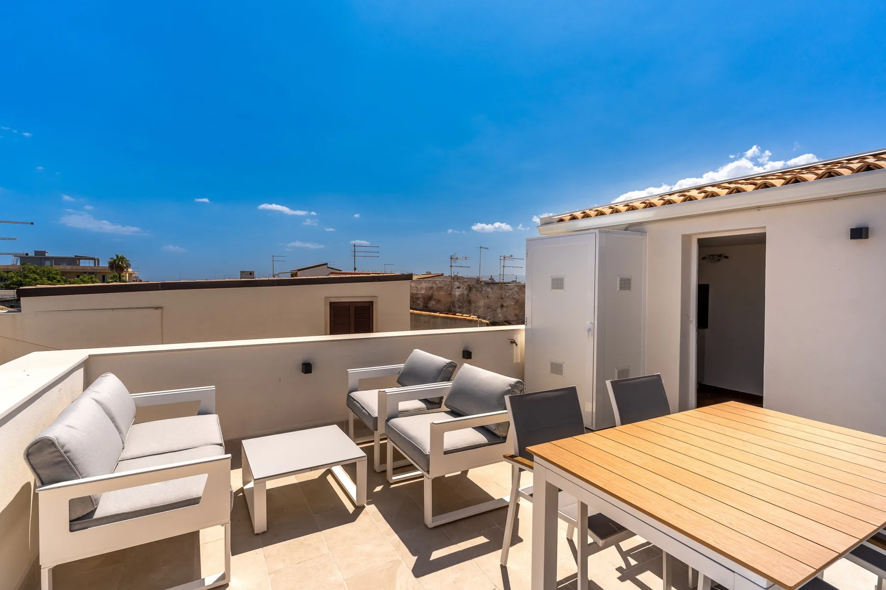 A rooftop terrace has outdoor seating and a wooden dining table beneath a blue sky.