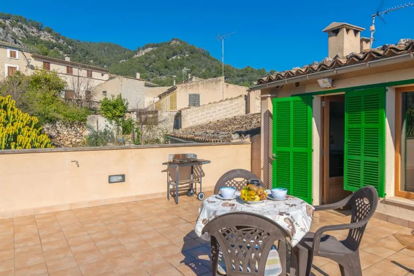 A sunny tiled terrace has outdoor seating and a table beside green shutters.