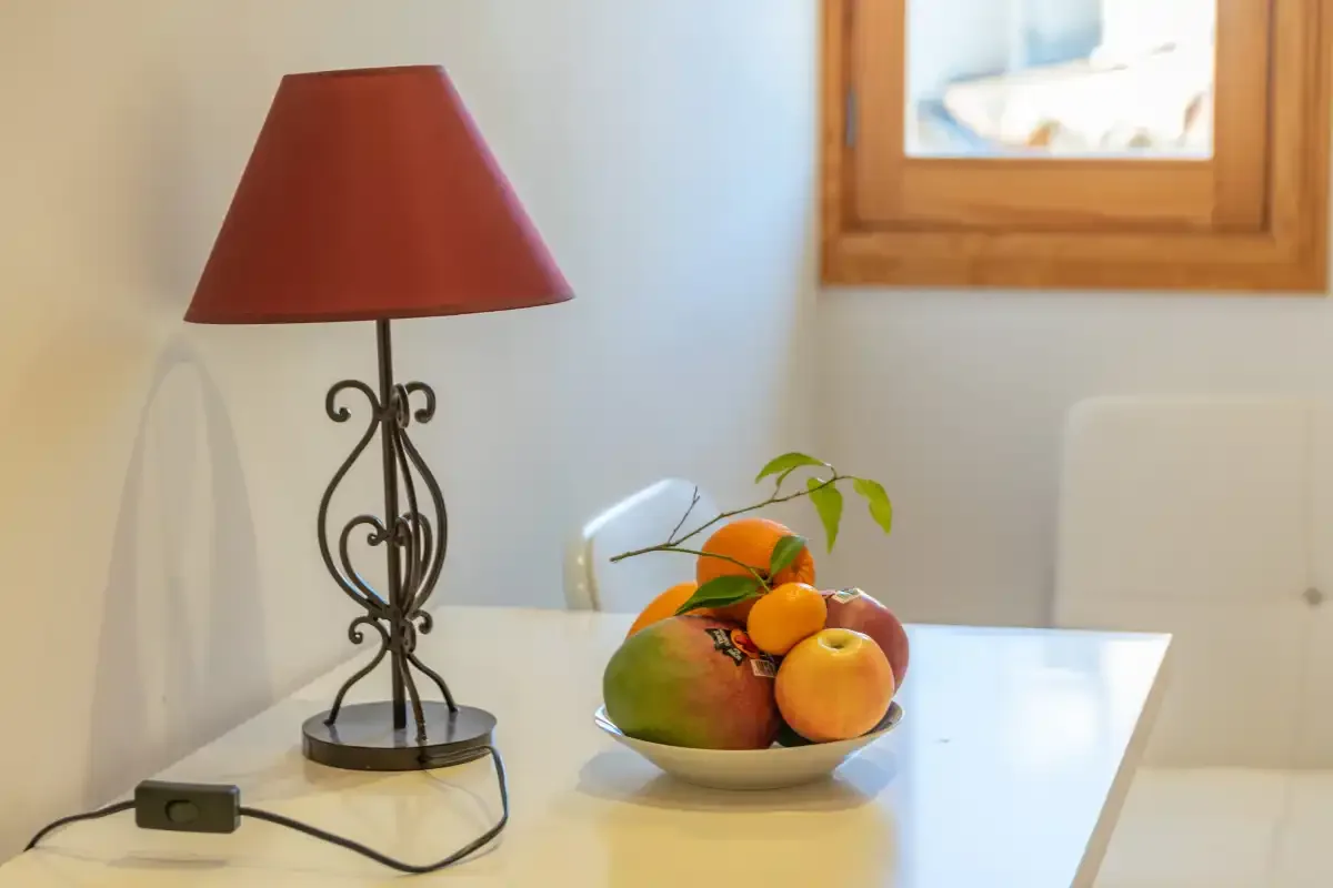 A dining table holds a fruit bowl beside a red-shaded lamp.