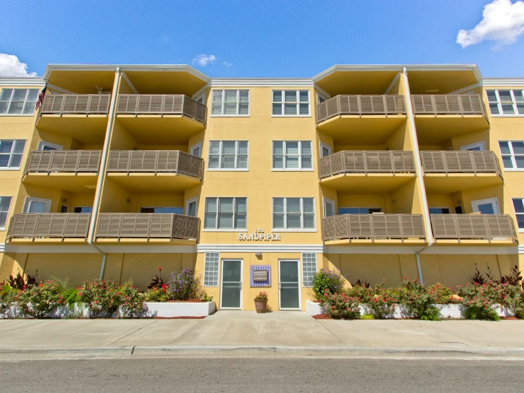 A yellow apartment building with balconies and flower beds along the entrance.