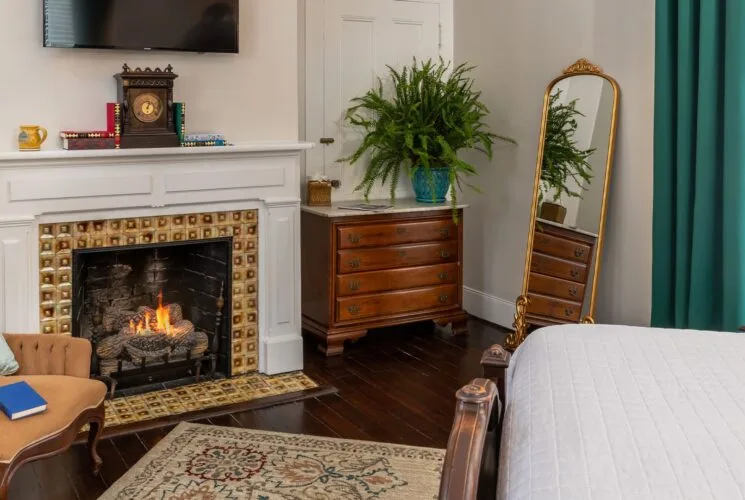 Bedroom with white walls, hardwood flooring, white bedding, fireplace, dresser, and antique armchair
