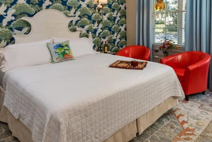 Bedroom with ornate wallpaper, hardwood flooring, white bedding, and a sitting area