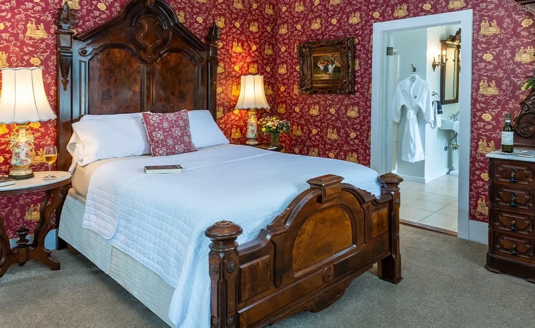 Bedroom with burgundy and gold ornate wallpaper, dark wooden bed and dresser, white bedding, and a view into the bathroom