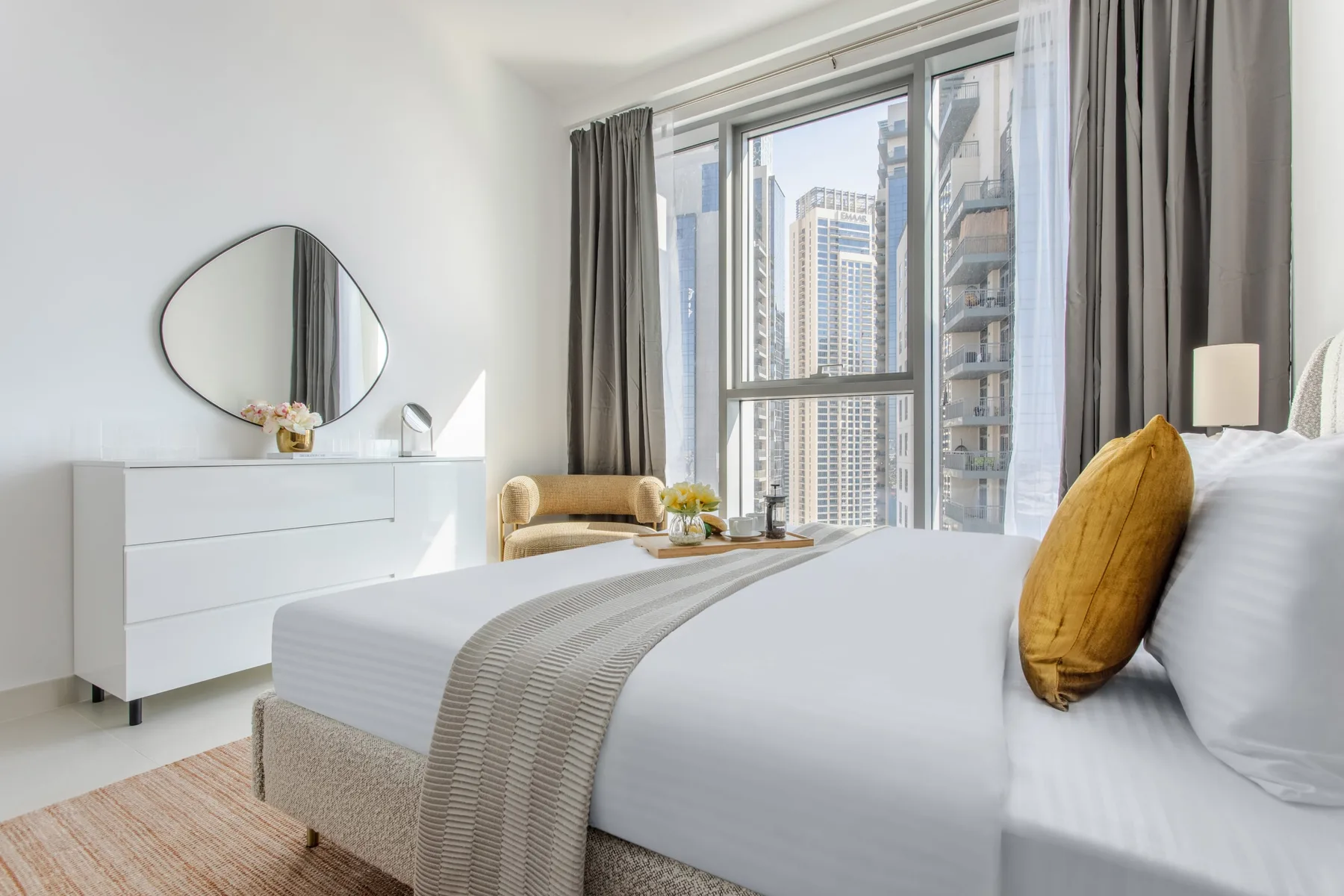 A bright bedroom with a neatly made bed beside large windows overlooking city buildings.