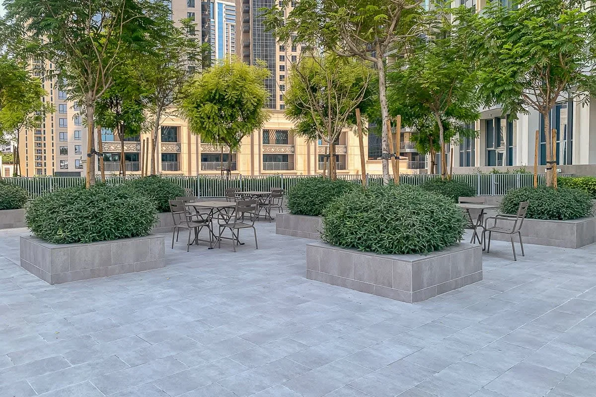 A paved courtyard has raised planting beds, trees, and outdoor seating.