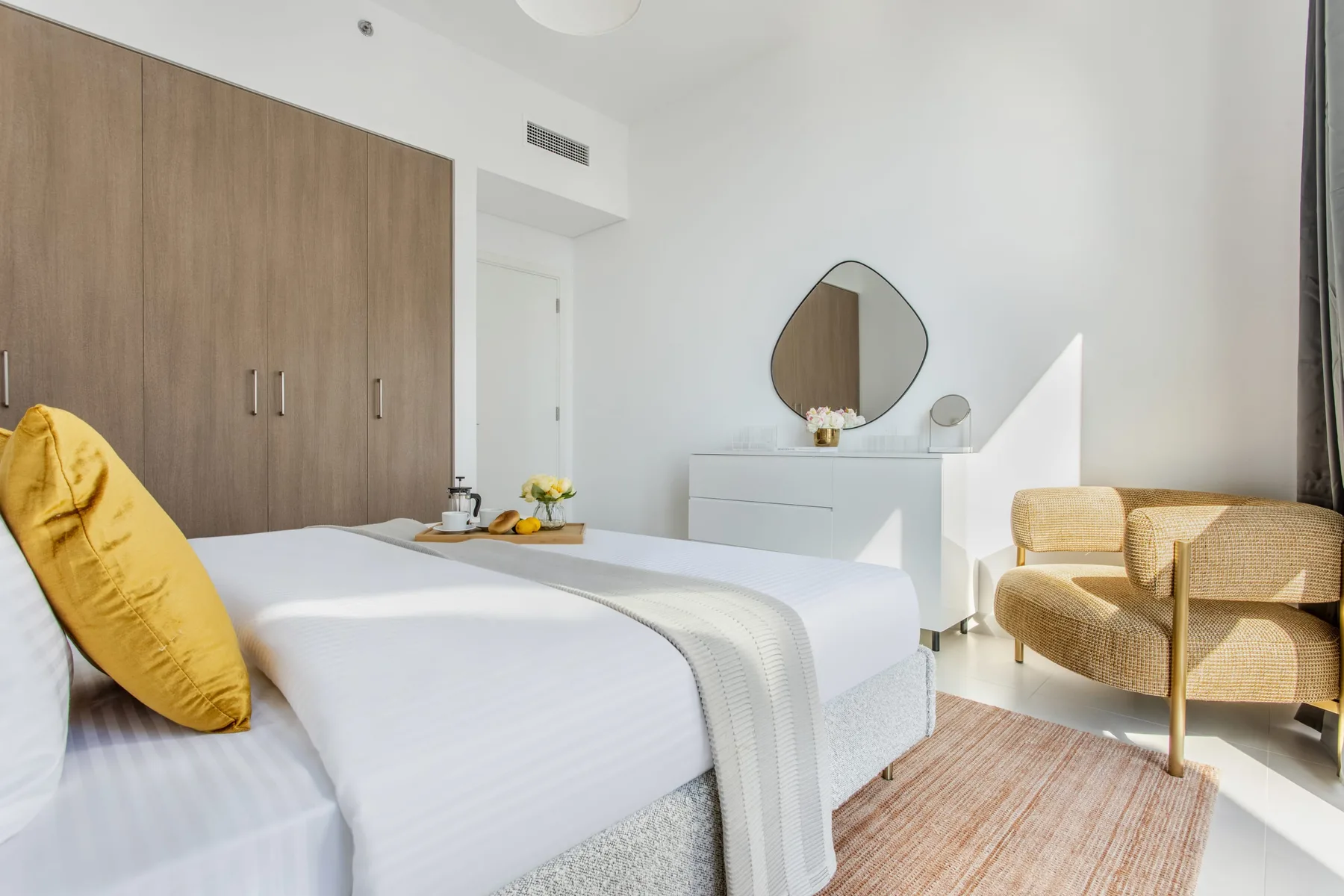 A bright bedroom with a neatly made bed, a wardrobe, and a vanity.