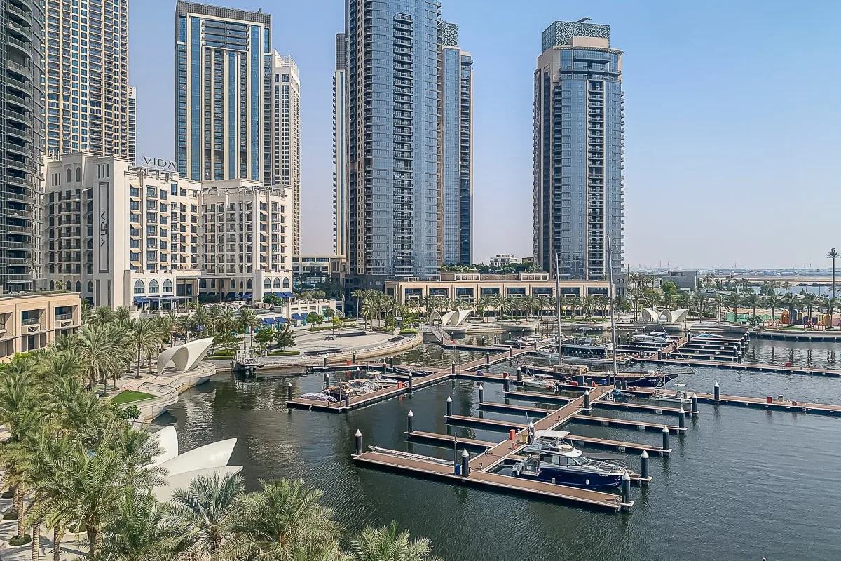 A waterfront marina with moored boats, palm trees, and tall buildings.