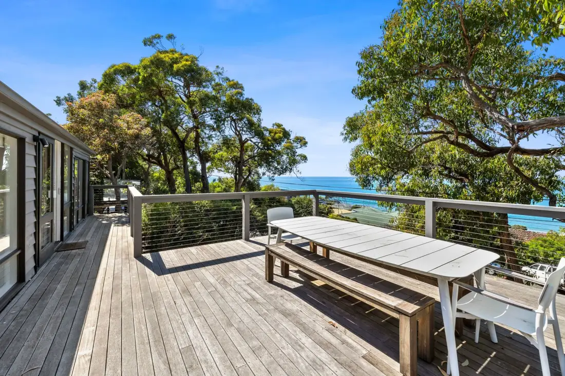 A wooden deck has outdoor dining furniture, leafy trees, and a view of the ocean.