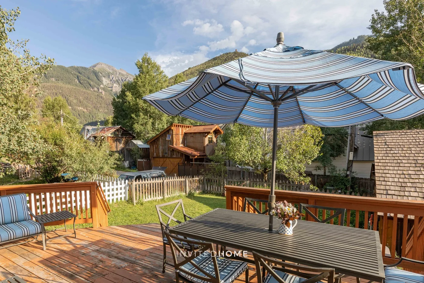 Zona de estar en una terraza bajo una sombrilla a rayas, con jardín cercado y montañas.